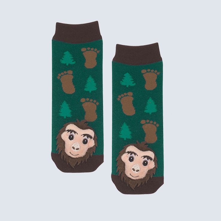 Two green socks against a white background. The socks feature a brown and green footprint motif and a plush Sasquatch charm on the toe.